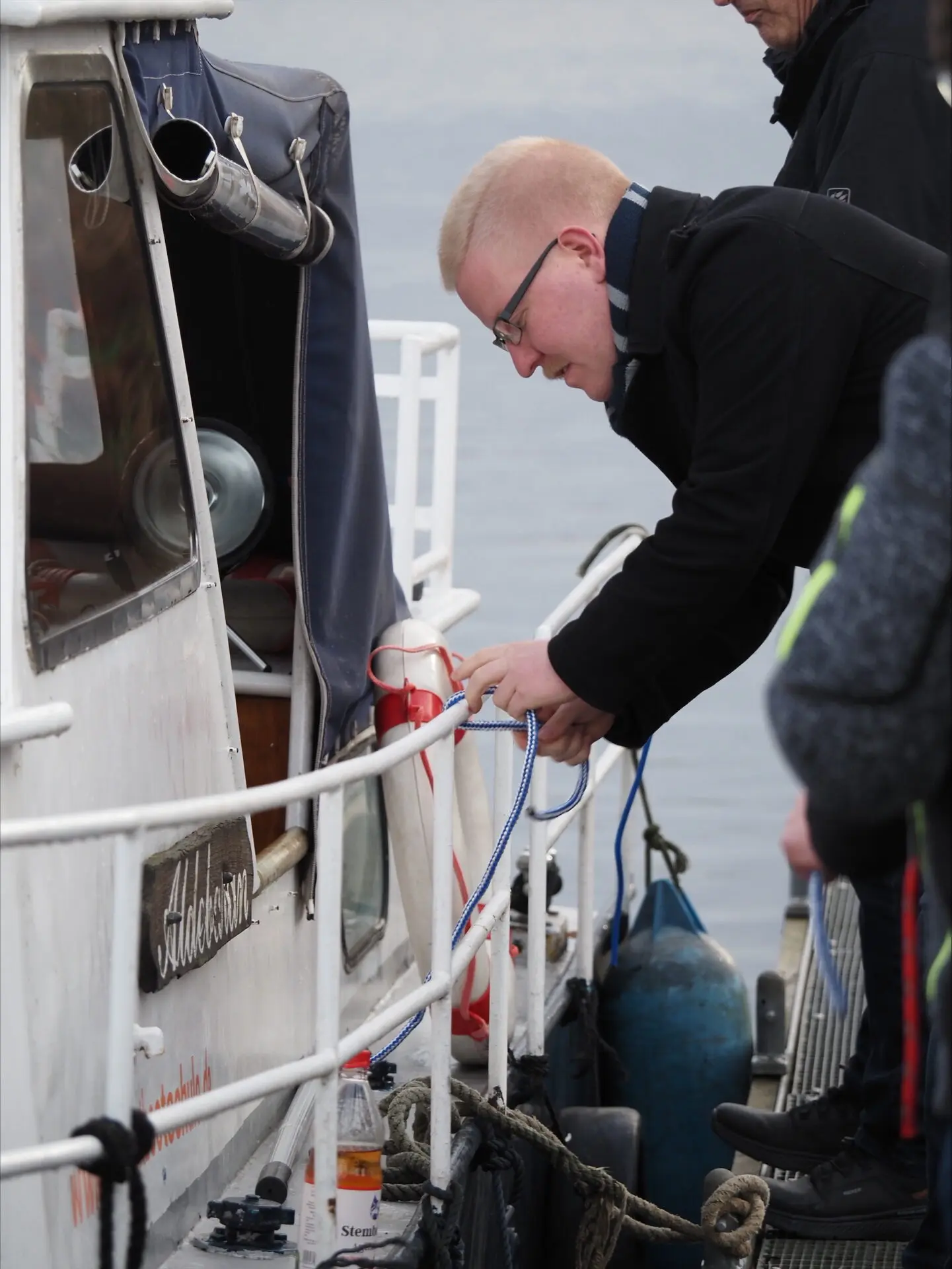 Junger Mann, der an einem Boot eine Leine befestigt, während andere Personen im Hintergrund stehen.