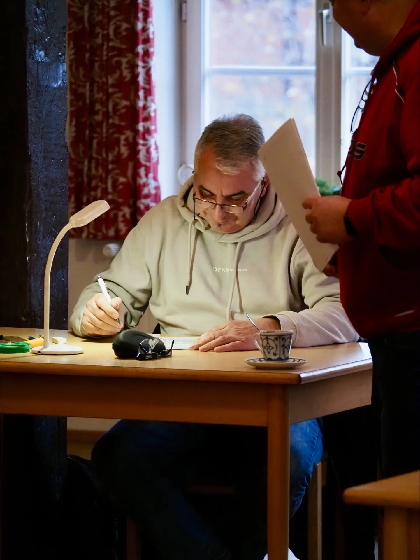 Mann mit Brille, der an einem Tisch sitzt und Notizen macht, während eine andere Person im Hintergrund steht.