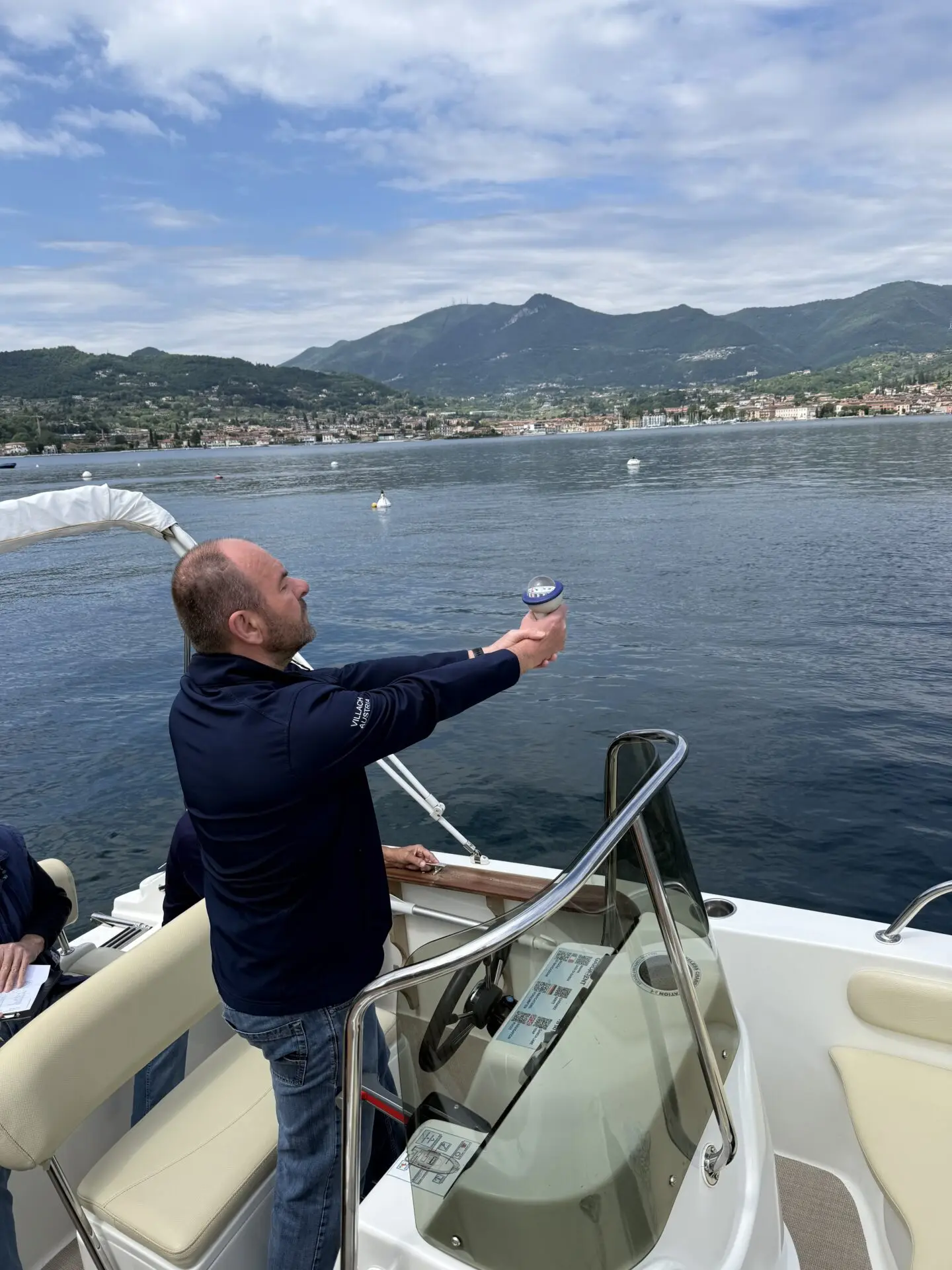 ab-sportbootschule_pruefung_30-04-2026_02 On Garda Lake, a man on a motorboat holds up a compass during a sailing test, with mountains, a lakeside town and cloudy sky behind him.