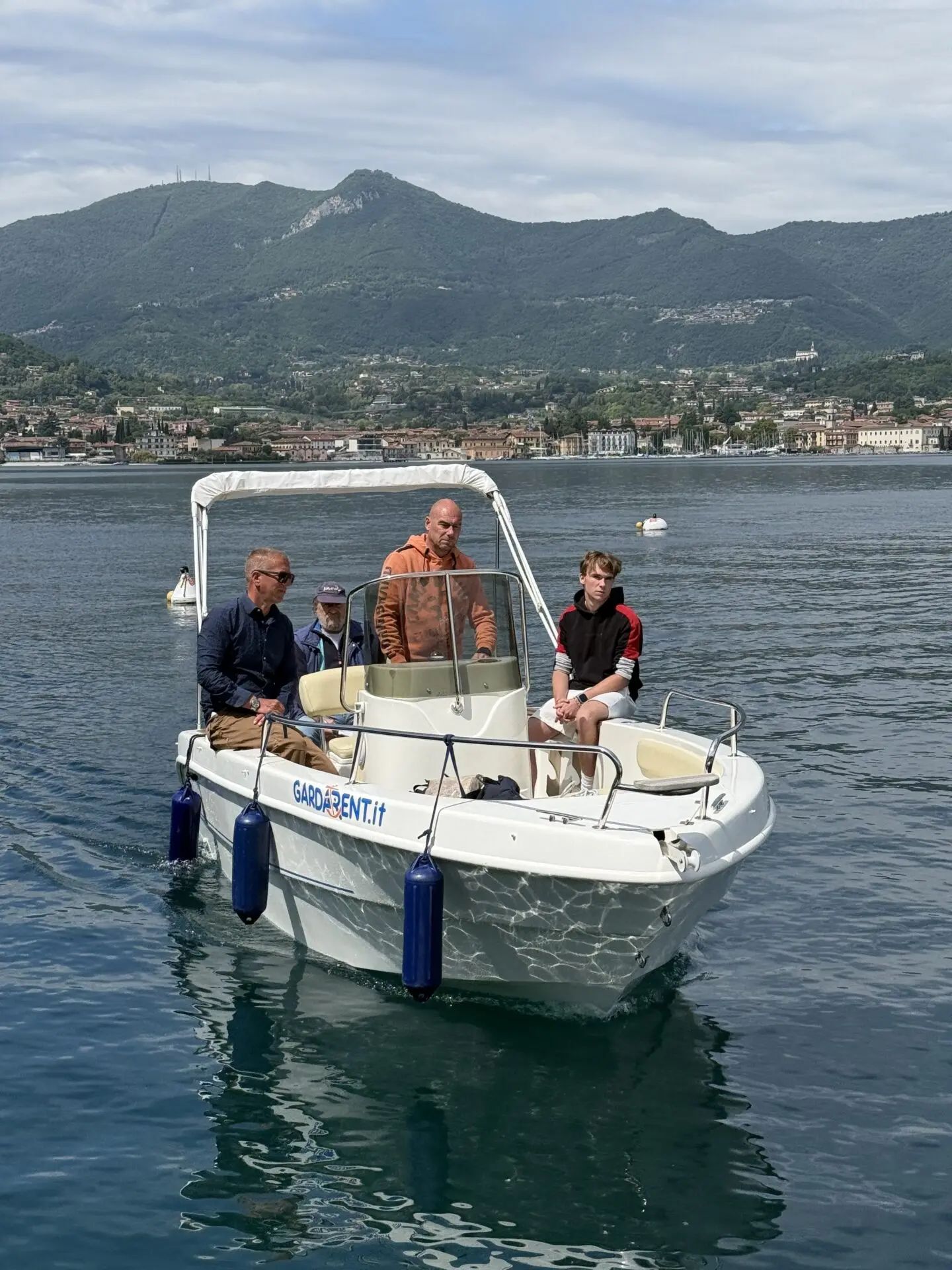 ab-sportbootschule_pruefung_30-04-2026_04 Auf dem Gardasee sitzt eine Gruppe auf einem Motorboot, während im Hintergrund die Uferstadt und Berge bei der Prüfung am Hotel Garden Zorzi sichtbar sind.