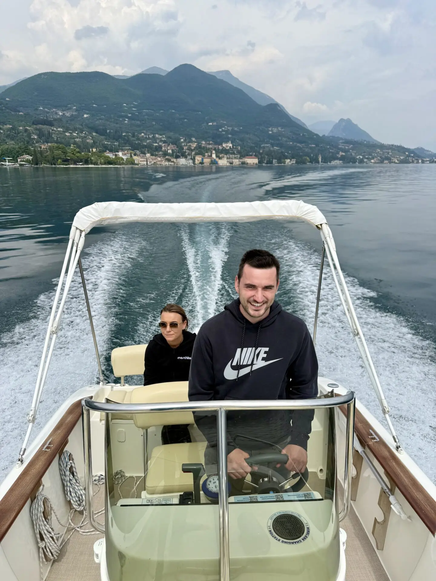 ab-sportbootschule_pruefung_30-04-2026_10 Zwei Personen steuern ein Motorboot über den Gardasee bei ruhigem Wasser, während sich im Hintergrund Berge und Uferorte abzeichnen.