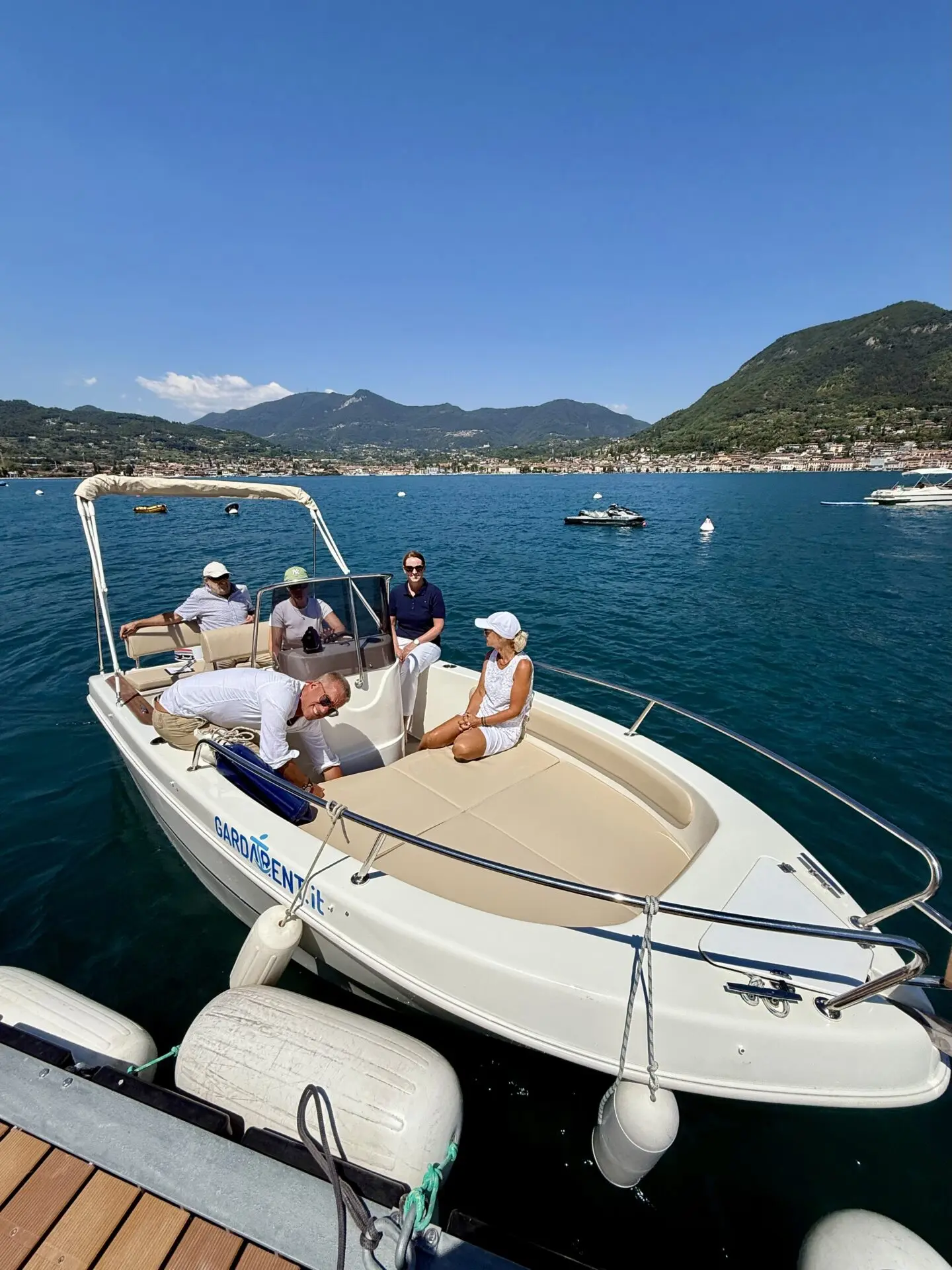 ab-sportbootschule_pruefung_09-07-2026_01 Mehrere Personen sitzen auf einem kleinen Motorboot auf dem tiefblauen See vor Bergen und einem Uferort.