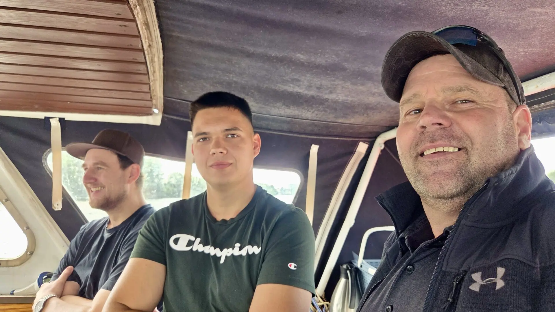 Drei Männer sitzen lächelnd in einem Boot, einer trägt ein Champion-Shirt und rechts sitzt ein Mann mit Kappe.