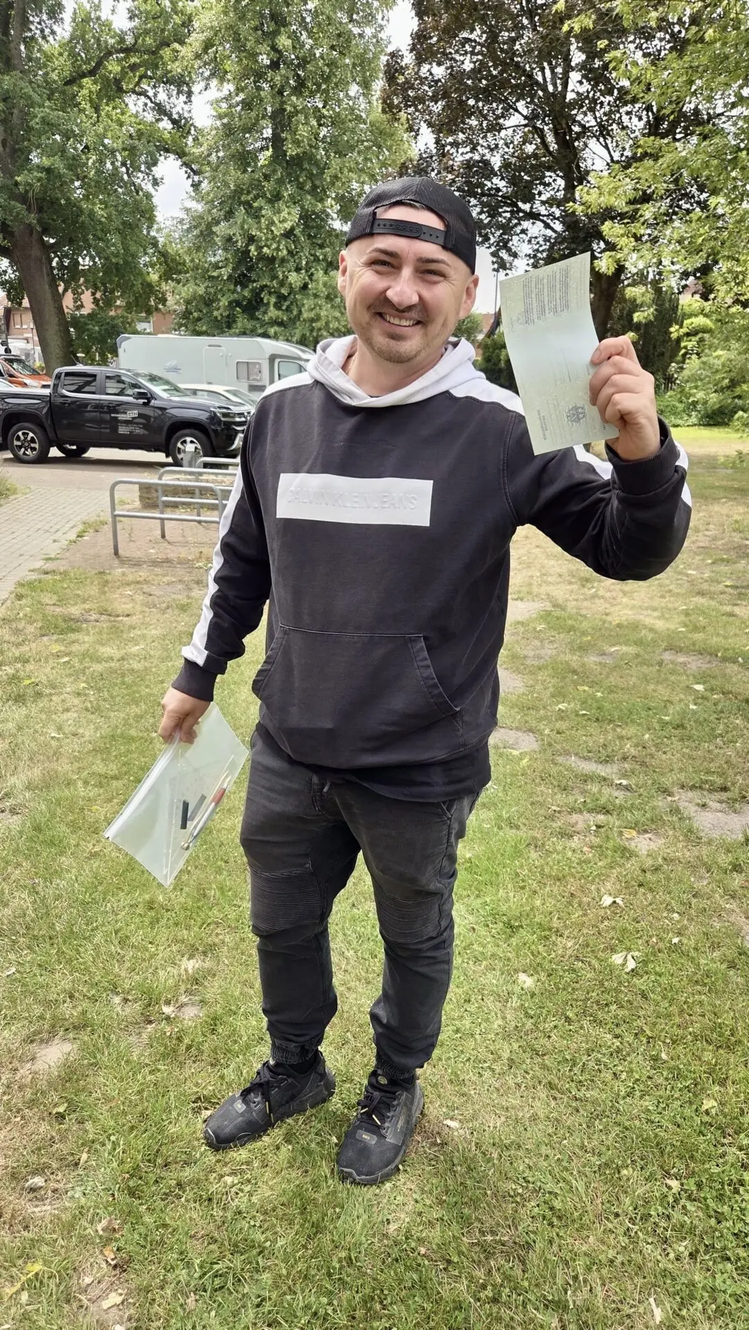 Ein lächelnder Mann im schwarzen Hoodie hält draußen auf einer Wiese Prüfungsunterlagen und einen Ausweis hoch.