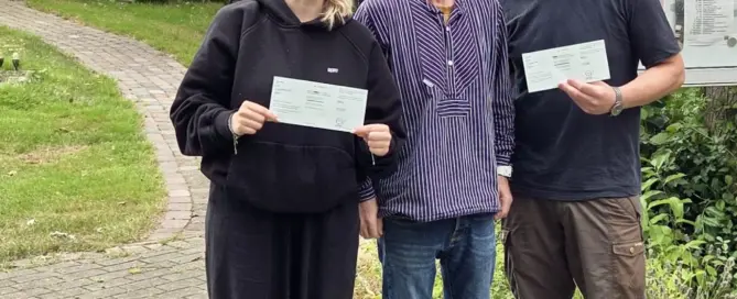 Zwei frischgebackene Prüflinge und ein Mann stehen vor Bäumen und halten ihre Prüfungsurkunden an der Weser.