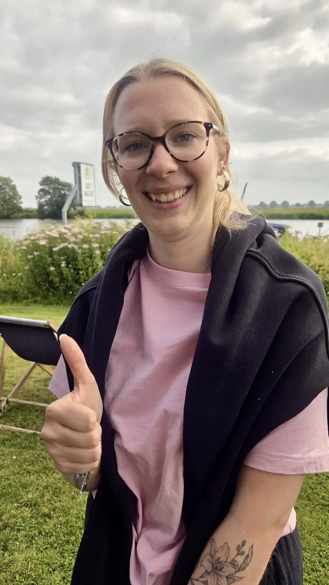 Eine lächelnde Frau mit Brille zeigt am Ufer der Weser den Daumen hoch und trägt ein rosa Shirt mit schwarzem Pullover.