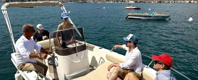 Mehrere Teilnehmer sitzen auf einem Motorboot auf dem Gardasee, während im Hintergrund Berge und weitere Boote zu sehen sind.