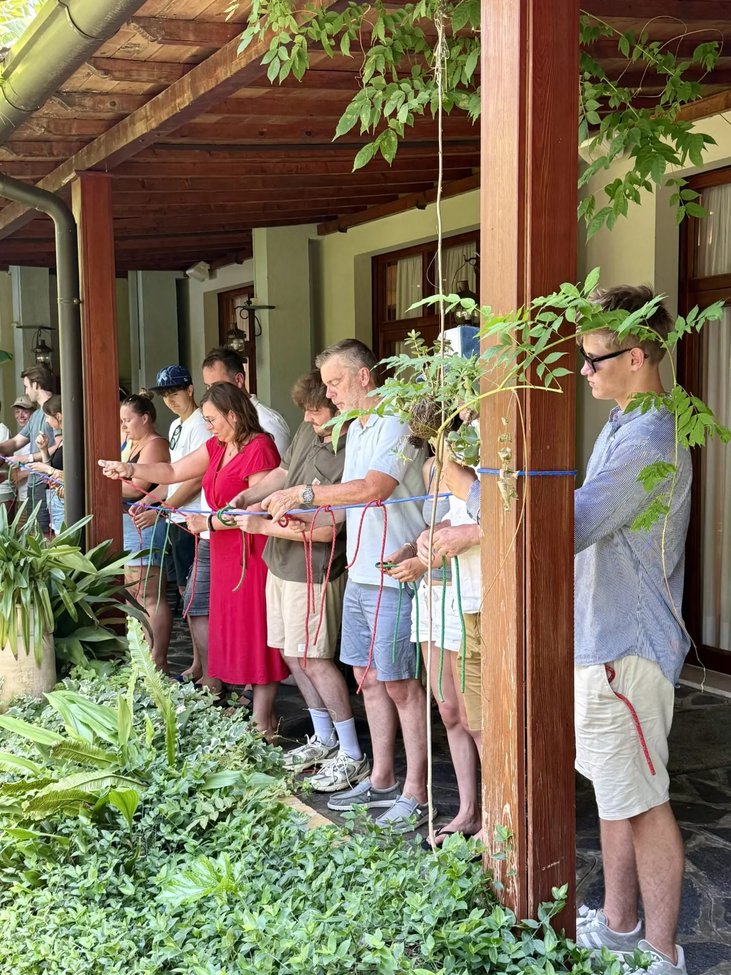 Mehrere Personen stehen auf einer überdachten Terrasse und halten farbige Seile, umgeben von üppigen Pflanzen.