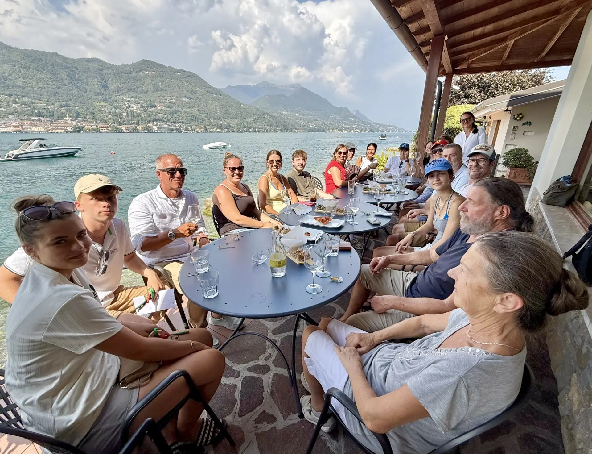 A group of boating students sits at lakeside tables on Lake Garda, with mountains and boats in the background.