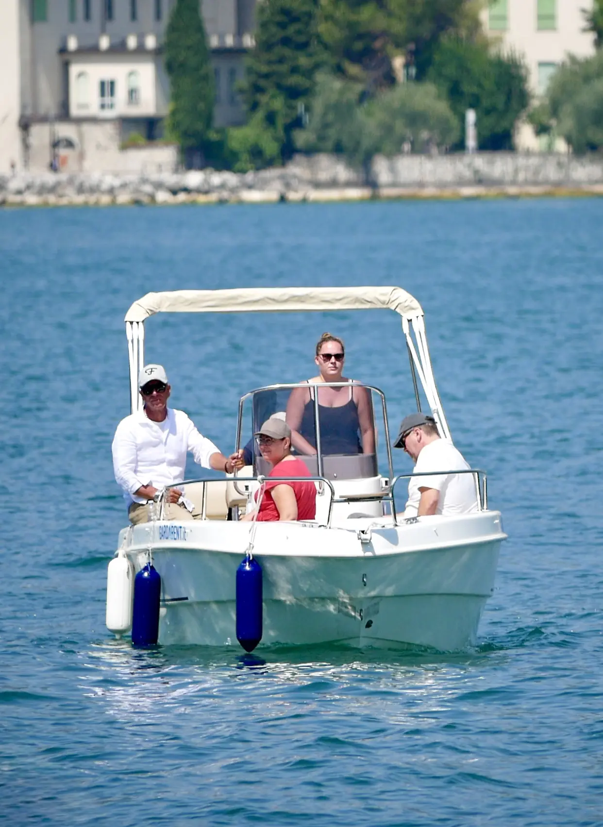 Vier Personen fahren mit einem kleinen Motorboot auf dem Gardasee, im Hintergrund liegt eine Uferstadt am Wasser.