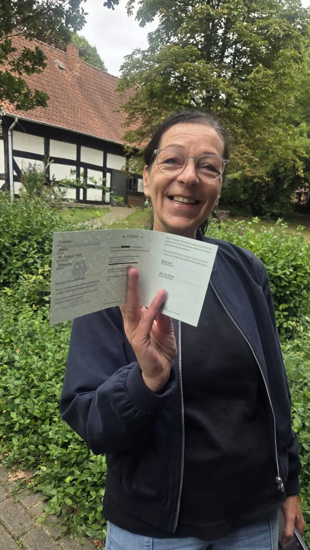 Eine lächelnde Frau hält ihre bestandene Sportbootführerschein-Prüfung vor einem historischen Fachwerkhaus an der Weser hoch.