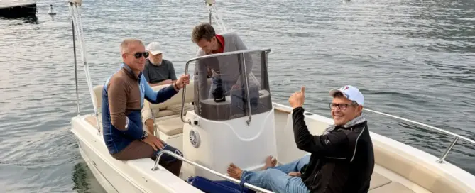 Vier Männer üben gemeinsam auf einem Motorboot auf dem Gardasee; San Felice del Benaco und die Alpen liegen am Ufer.