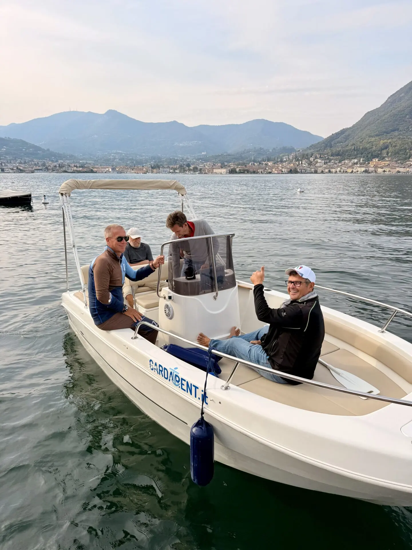 Vier Männer üben gemeinsam auf einem Motorboot auf dem Gardasee; San Felice del Benaco und die Alpen liegen am Ufer.